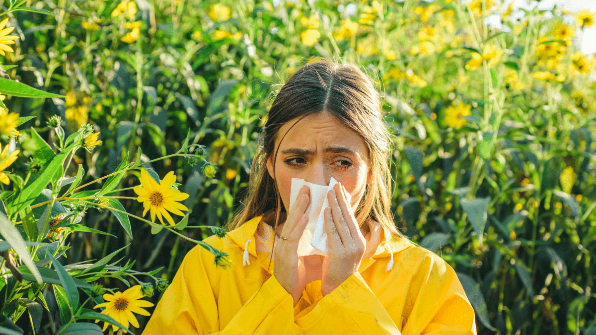 A woman trying to manage summer allergies outdoors sneezes while covering her mouth with a tissue.