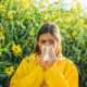 A woman trying to manage summer allergies outdoors sneezes while covering her mouth with a tissue.