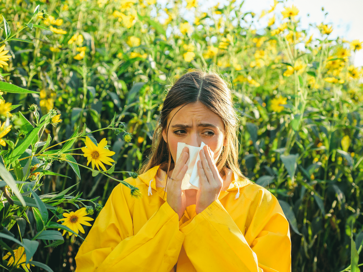 A woman trying to manage summer allergies outdoors sneezes while covering her mouth with a tissue.
